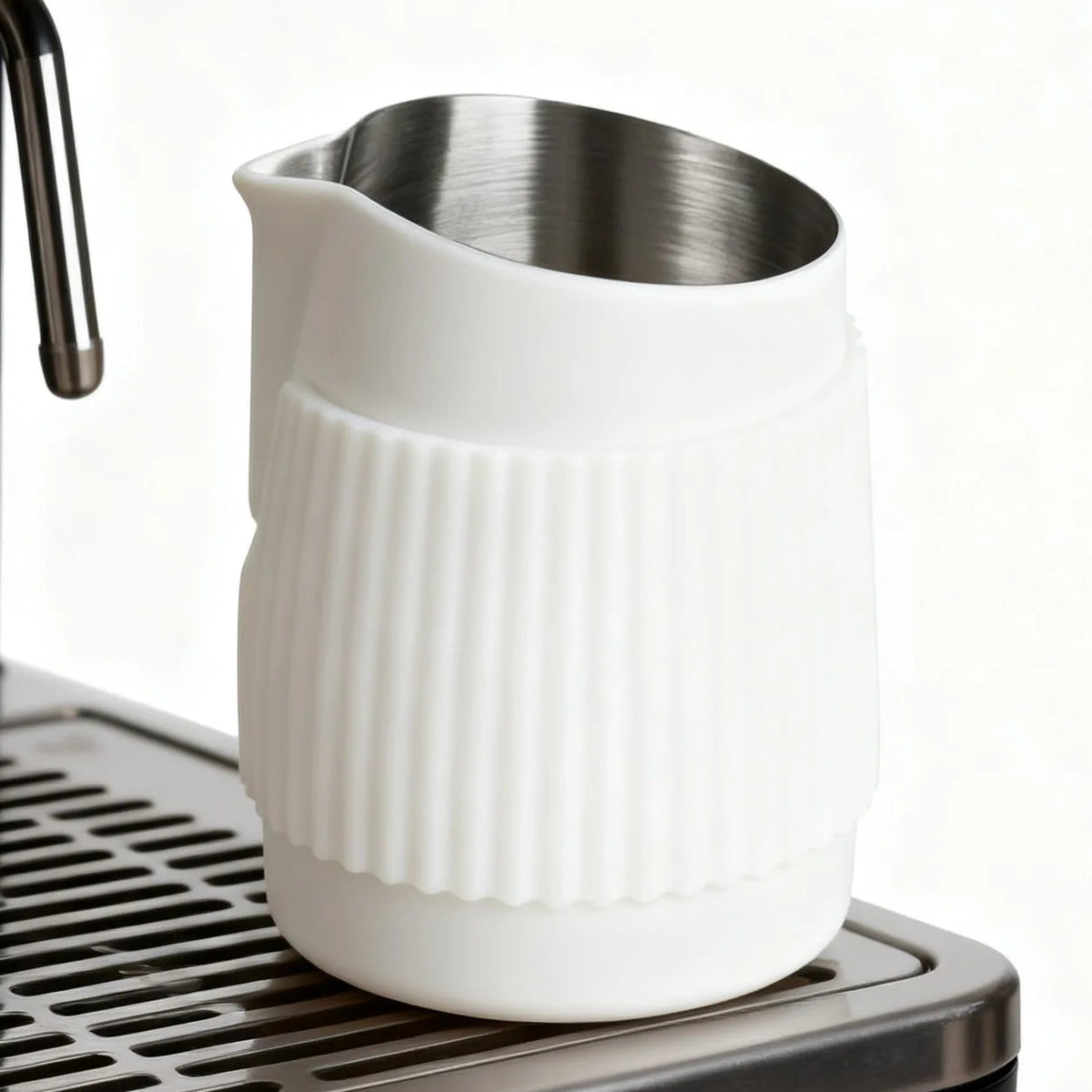 A white ceramic milk pitcher with a ribbed texture and a stainless steel interior, shown resting on the drip tray of a black espresso machine against a white background.
