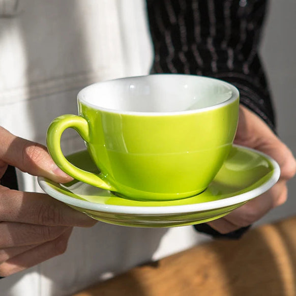 A bright lime green ceramic espresso cup with a matching saucer, held by hands wearing a white apron and a black pinstriped jacket. The scene is lit by warm, direct sunlight casting shadows.