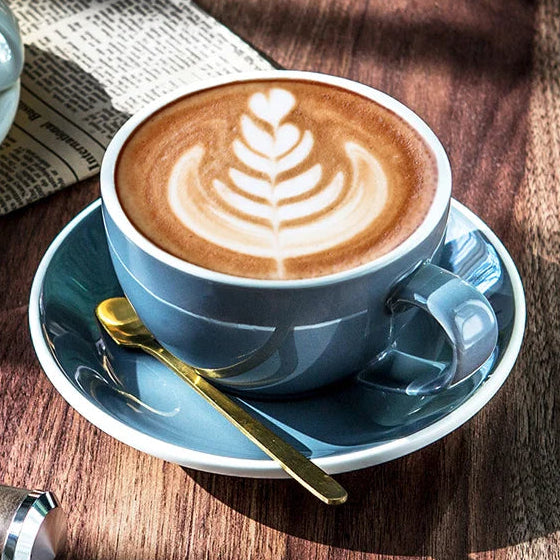 A cup of latte coffee with leaf-shaped latte art in a dusty blue ceramic mug on a matching saucer, set on a wooden table with a gold spoon, newspaper, and another cup in the background.