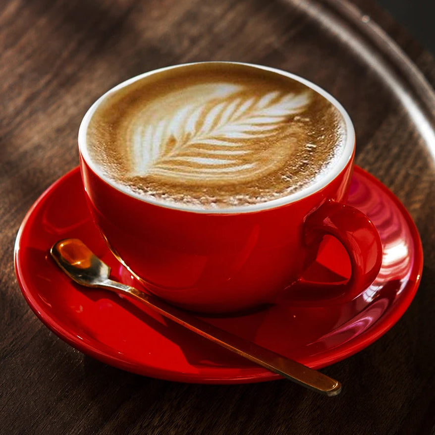 A red ceramic coffee cup filled with latte art sits on a matching red saucer with a gold spoon, placed on a dark wooden tray.