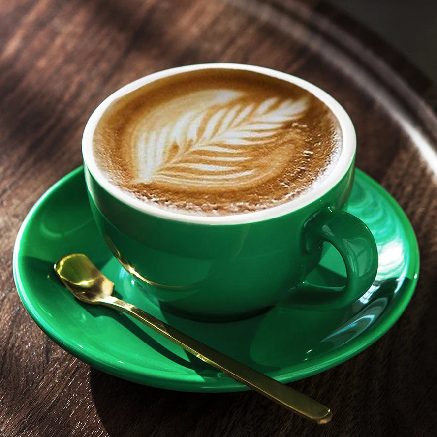 A green ceramic coffee cup filled with latte art sits on a matching green saucer with a gold spoon, placed on a dark wooden table in warm sunlight.