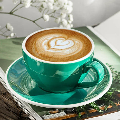 A teal ceramic coffee cup filled with latte art sits on a matching saucer, placed on top of an open magazine on a rustic wooden surface with white flowers in the background.