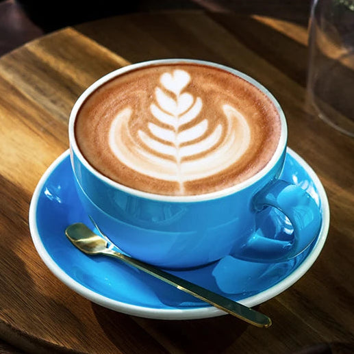 A bright blue ceramic coffee cup and saucer set holding a latte with leaf latte art, styled on a round wooden board with a small spoon, flowers in a glass jar, and a dark mug in a warm indoor setting.