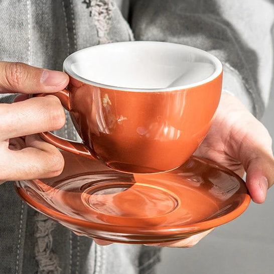 A person in a distressed gray denim jacket holds a glossy terracotta orange ceramic espresso cup with a matching saucer. The cup has a white interior, and the background is a soft, out-of-focus gray.