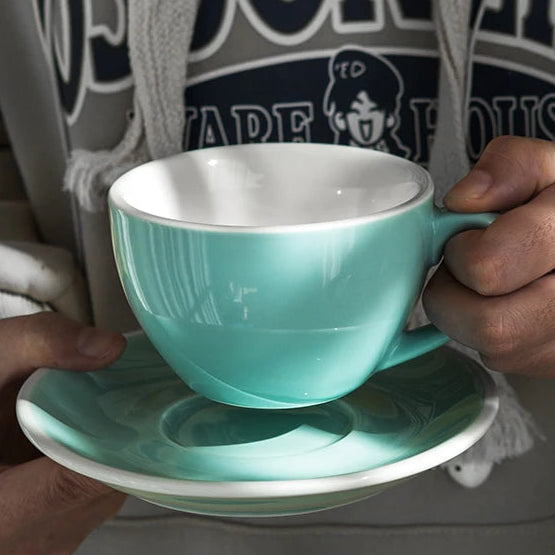 A person in a gray hoodie holds a mint green ceramic teacup and matching saucer. The hoodie has a visible 'Bonneville' graphic on the chest.