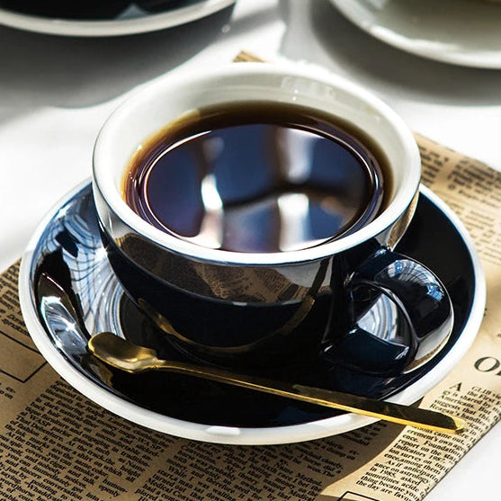 A dark navy blue ceramic coffee cup and saucer set on a newspaper mat, with a gold spoon resting on the saucer. A matching cup and saucer are blurred in the background, along with a slice of cake and small white flowers.
