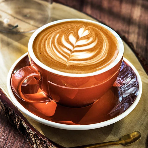 A cup of latte coffee with leaf latte art in a terracotta-colored ceramic cup and saucer, set on a wooden slice tray with a spoon and a glass vase in the background.