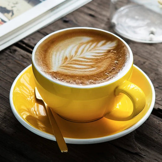 A bright yellow ceramic coffee cup filled with latte art sits on a matching yellow-and-white saucer with a gold spoon, set on a rustic dark wooden table. An open magazine and small glass vase with white flowers are blurred in the background.
