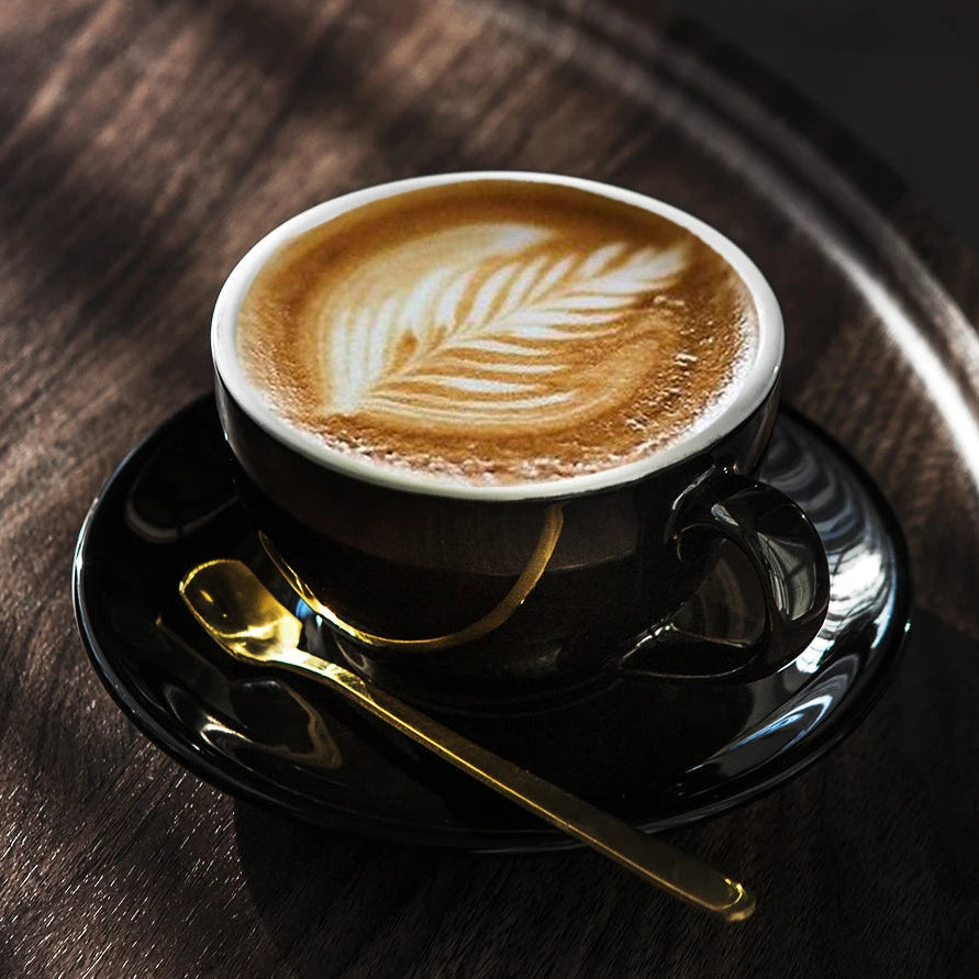 A black coffee cup with a white interior, filled with latte art, sits on a matching black saucer with a gold spoon on a dark wooden table.