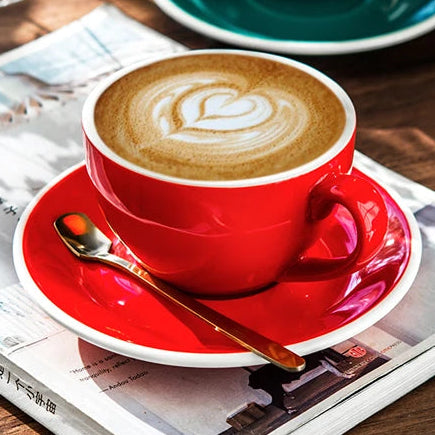 A red ceramic coffee cup with a matching saucer sits on a wooden table, filled with latte macchiato and topped with heart-shaped foam. Another teal cup and saucer are visible in the background, along with a magazine, a spoon, and small white flowers in a glass vase.