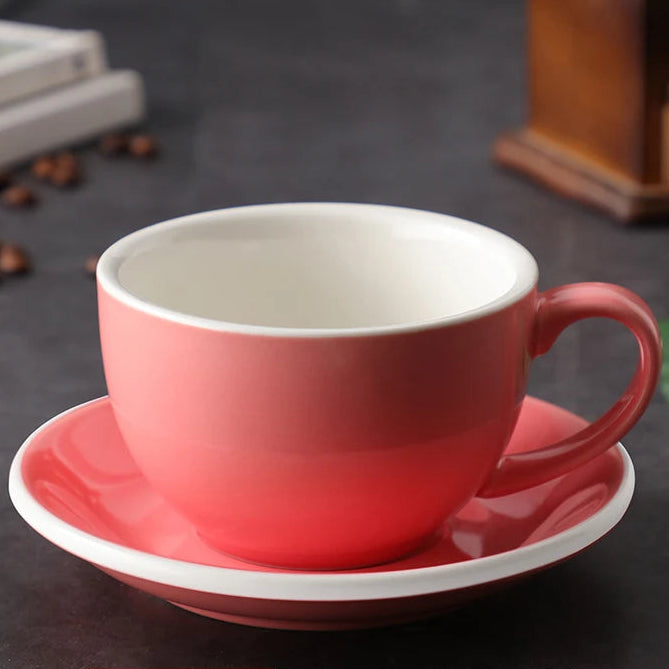 A pink ceramic coffee cup with a matching saucer, featuring a white interior and rim. The cup and saucer are displayed on a dark tabletop with coffee beans and blurred background props.