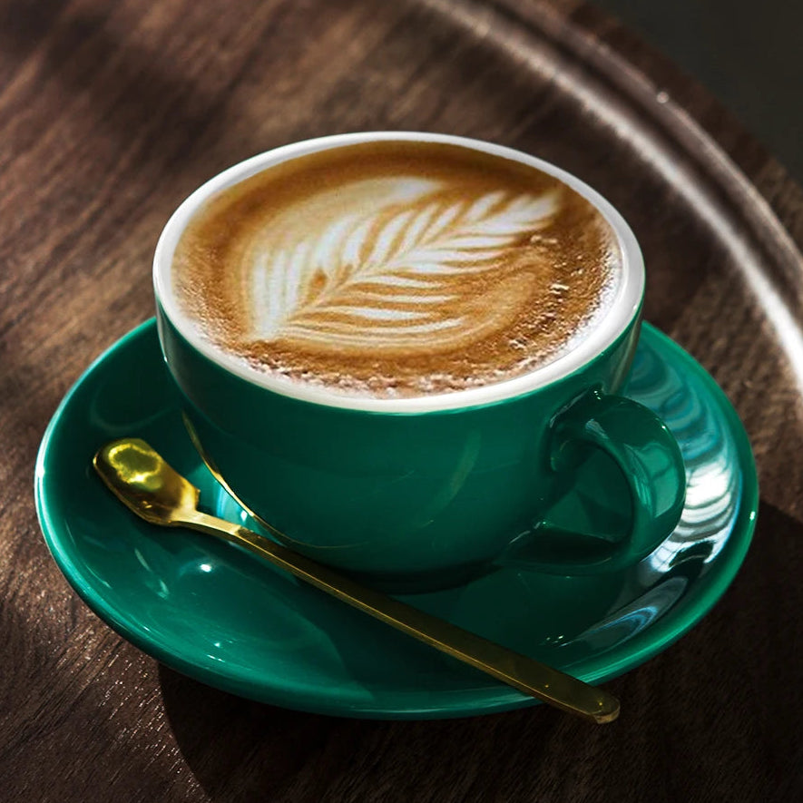 A green ceramic coffee cup filled with latte art sits on a matching green saucer with a gold spoon, placed on a dark wooden tray.