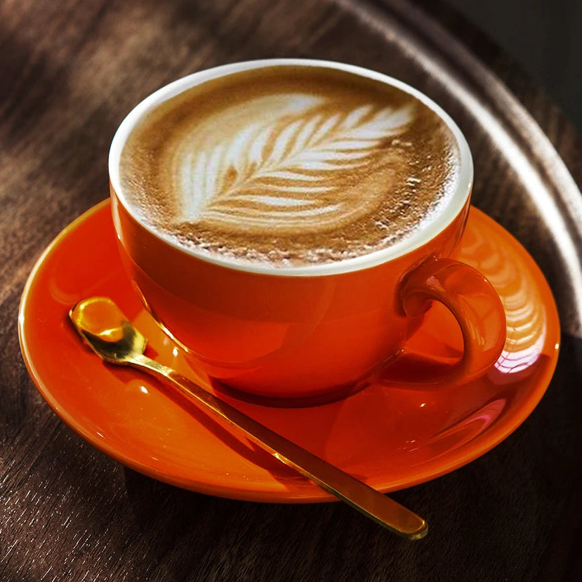 An orange ceramic coffee cup filled with latte art sits on a matching orange saucer with a gold spoon, placed on a dark wooden tray.