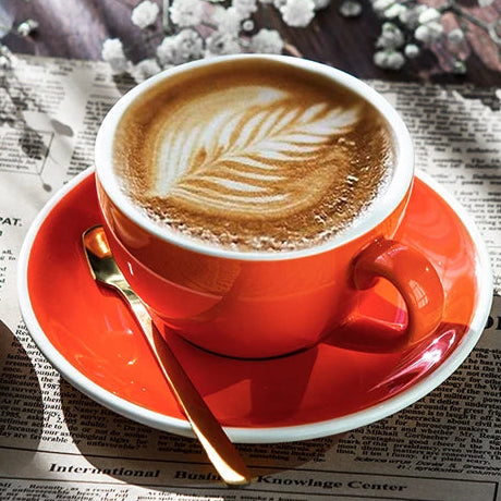 A warm orange ceramic coffee cup filled with latte art sits on a matching saucer, placed on top of newspaper with scattered white flowers around it. A second yellow cup and saucer are partially visible at the left edge.